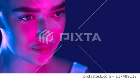 A woman face is brightly lit up with a combination of vibrant blue and pink lights, creating a striking contrast against her features. The blue and pink hues interplay on her skin, copy space 117499112