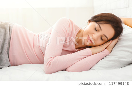 A young woman enjoys a restful sleep on a comfortable bed with sunlight streaming through the window, creating a serene atmosphere. 117499148