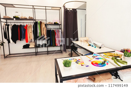 A room cluttered with various clothing items strewn across the floor and hanging on racks. A table in the center is piled high with more clothes, creating a chaotic and untidy environment. 117499149