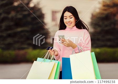 Korean woman standing holding multiple shopping bags in one hand while using a cell phone in the other. She appears busy and focused on her phone amidst her shopping activities. Korean woman standing holding multiple shopping bags in one hand while using a cell phone in the other. She appears busy and focused on her phone amidst her shopping activities. 117499238