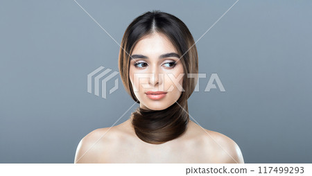 A young woman with long, smooth brown hair wrapped stylishly around her neck poses against a plain gray backdrop. Her expression is serene and thoughtful. A young woman with long, smooth brown hair wrapped stylishly around her neck poses against a plain gray backdrop. Her expression is serene and thoughtful. 117499293