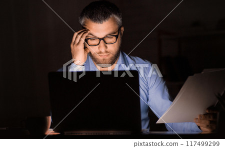 A man in glasses is seen working late in a dimly lit office. He holds documents in one hand while looking intently at a laptop screen. The setting suggests diligent effort and focus 117499299