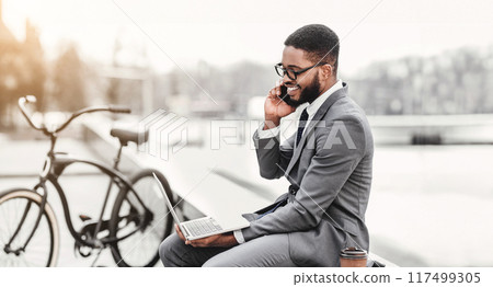 Black man professional is using his laptop and talking on his phone while sitting outside, with a bike beside him. The scene is modern and energetic, showcasing multitasking in a fresh setting. Black man professional is using his laptop and talking on his phone while sitting outside, with a bike beside him. The scene is modern and energetic, showcasing multitasking in a fresh setting. 117499305