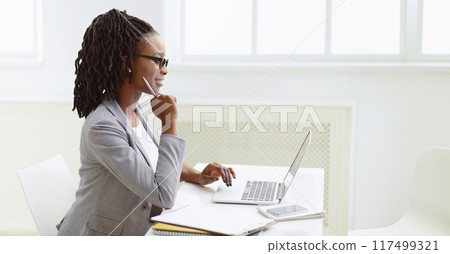 African American woman in glasses is typing on her laptop at an office desk. The setting is bright and professional, with a clean desk and modern decor, indicating concentration. African American woman in glasses is typing on her laptop at an office desk. The setting is bright and professional, with a clean desk and modern decor, indicating concentration. 117499321