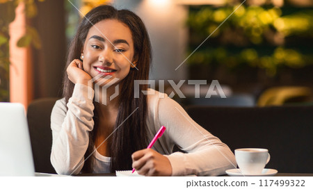 African American young girl is sitting at a table, working on her laptop with a pen in hand. She appears focused and engaged in her work, surrounded by a simple setting. African American young girl is sitting at a table, working on her laptop with a pen in hand. She appears focused and engaged in her work, surrounded by a simple setting. 117499322