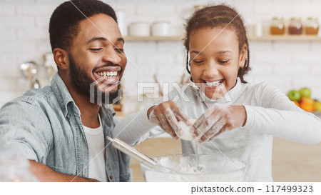 Cute african girl adding eggs into dough bowl, father teaching daughter cooking in kitchen 117499323