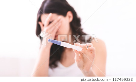 A young woman is holding a pregnancy test in her hand while covering her face with her other hand. She appears to be emotional, possibly in shock or disbelief. A young woman is holding a pregnancy test in her hand while covering her face with her other hand. She appears to be emotional, possibly in shock or disbelief. 117499328