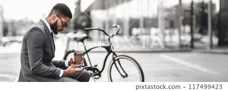 African American businessman holding coffee cup and phone, sitting near a bicycle in urban setting, panorama with copy space African American businessman holding coffee cup and phone, sitting near a bicycle in urban setting, panorama with copy space 117499423