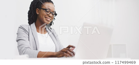 An office employee black lady is focused on her computer at her desk. The atmosphere is professional and modern, with a clean desk and bright space, highlighting concentration. 117499469