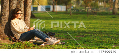 Asian young woman enjoys a peaceful afternoon in the park, sitting against a tree, and checking her phone. She has books next to her, studying or simply enjoying some leisure time outdoors, copy space 117499519