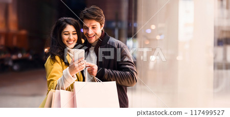 Happy couple ordering taxi on cellphone, walking near city center after shopping on weekend 117499527