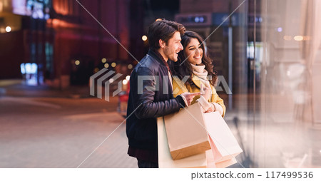 Window shopping. Young couple looking at showcase, walking near city center in the evening, free space Window shopping. Young couple looking at showcase, walking near city center in the evening, free space 117499536