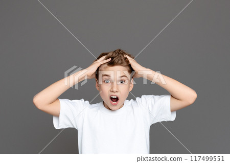 A shocked teen boy is captured with wide eyes and hands clutching his head against an isolated grey backdrop, suggesting alarm A shocked teen boy is captured with wide eyes and hands clutching his head against an isolated grey backdrop, suggesting alarm 117499551