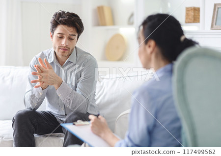 Unhappy man sitting on a couch engaged in conversation with a therapist in a office setting. The man appears focused and attentive while the therapist listens and provides guidance. 117499562