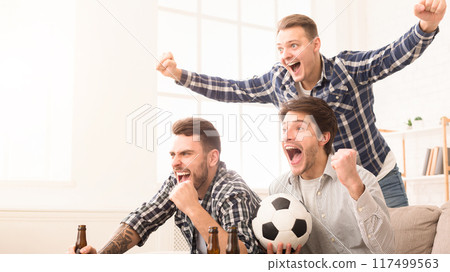 Three enthusiastic men celebrate with cheers and raised hands, holding a soccer ball and beer bottles 117499563