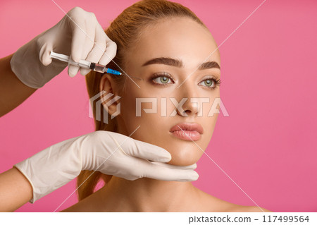 Cosmetology concept. Young woman getting beauty injection near eyes, pink background Cosmetology concept. Young woman getting beauty injection near eyes, pink background 117499564