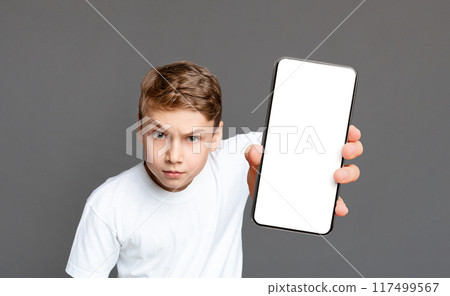 A young boy with a serious expression holds out a smartphone with a blank screen towards the camera on a gray background A young boy with a serious expression holds out a smartphone with a blank screen towards the camera on a gray background 117499567