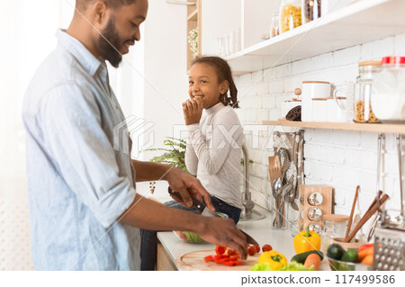 Black Man Making Food With His Sweet Daughter In Kitchen, cutting vegetables, girl eating cookie and smiling, side view, copy space Black Man Making Food With His Sweet Daughter In Kitchen, cutting vegetables, girl eating cookie and smiling, side view, copy space 117499586