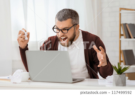 A bearded man with glasses is sitting in a bright office, visibly frustrated and shouting at his laptop. Papers and a plant are on the desk, suggesting a working environment. A bearded man with glasses is sitting in a bright office, visibly frustrated and shouting at his laptop. Papers and a plant are on the desk, suggesting a working environment. 117499592