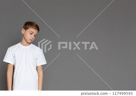 A sad teenage boy gazing downwards, expressing despondency He stands isolated with a grey background, copy space A sad teenage boy gazing downwards, expressing despondency He stands isolated with a grey background, copy space 117499593