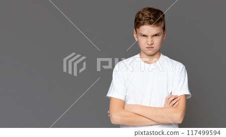 A young boy stands with his arms crossed, frowning against a solid gray background, displaying a look of dissatisfaction or disagreement A young boy stands with his arms crossed, frowning against a solid gray background, displaying a look of dissatisfaction or disagreement 117499594