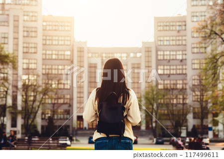 A woman stands in front of a towering building, looking up at its impressive height. The sleek architecture and glass facade of the building contrast against the sky, back view A woman stands in front of a towering building, looking up at its impressive height. The sleek architecture and glass facade of the building contrast against the sky, back view 117499611