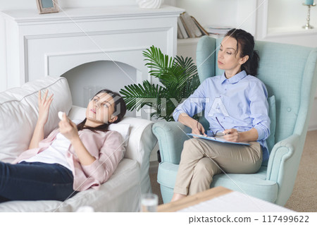 Emotional woman is lying on a couch while talking to a therapist seated nearby. The setting appears calm and welcoming, suggesting a home office environment. 117499622
