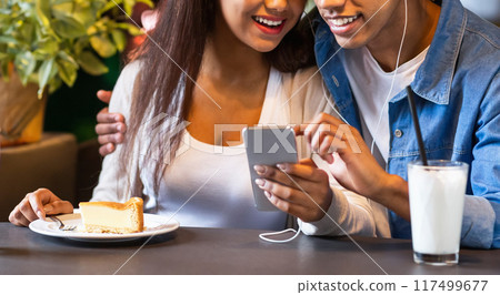 A young black couple is seated at a cafe table, smiling and listening to music through a shared set of earphones, looking at a smartphone together, cropped A young black couple is seated at a cafe table, smiling and listening to music through a shared set of earphones, looking at a smartphone together, cropped 117499677