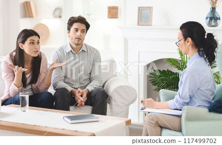 Emotional man and a woman are sitting on a couch, engaged in conversation with a therapist. The therapist is listening attentively and offering guidance in a professional setting. 117499702