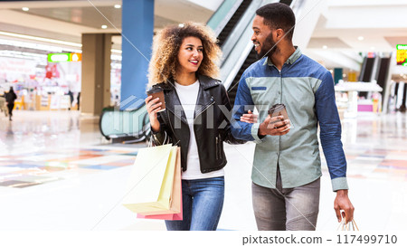 A happy African American couple is walking through a shopping mall, engaged in a lively conversation. They are casually dressed and carry multiple shopping bags, drinking takeaway coffee 117499710