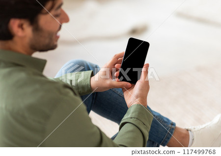 A man, wearing a green shirt and blue jeans, is sitting on a couch and holding a smartphone in his hands. The screen of the phone is black, and the man is looking at the phone 117499768