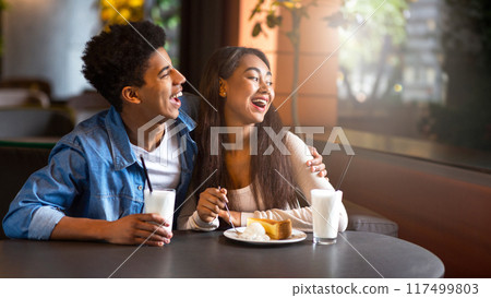 A young black couple sits together at a cozy cafe, smiling and laughing while enjoying breakfast. Both are holding glasses of milk, and a plate with food is in front of them, copy space A young black couple sits together at a cozy cafe, smiling and laughing while enjoying breakfast. Both are holding glasses of milk, and a plate with food is in front of them, copy space 117499803