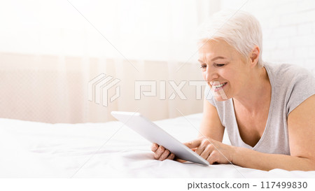 Happy senior woman lying in bed, smiling and watching pictures of her grandchildren on digital tablet, panorama with empty space Happy senior woman lying in bed, smiling and watching pictures of her grandchildren on digital tablet, panorama with empty space 117499830