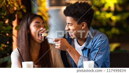 African American guy and girl are seated at a table in a restaurant, enjoying ice cream. They are both holding cones and actively eating. The restaurant setting is casual and comfortable. 117499834