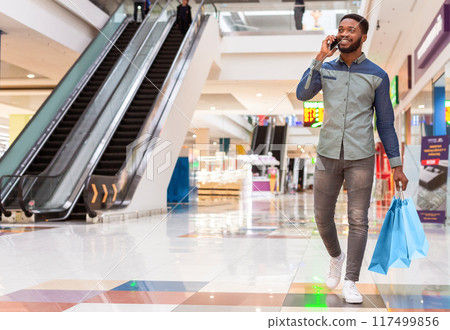 A black man is seen walking through a bustling shopping mall, engaged in a phone conversation. He appears focused on his call amidst the hustle and bustle of the malls activity, copy space 117499856