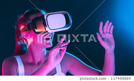 A woman is depicted wearing a virtual reality headset, fully immersed in a digital environment. The headset covers her eyes, ears, and head, indicating her engagement with the virtual world. 117499864