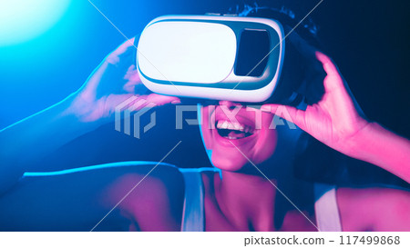 A woman is wearing a virtual reality headset, fully immersed in a digital environment. The headset covers her eyes and ears as she interacts with the virtual world displayed in the headsets lenses. 117499868