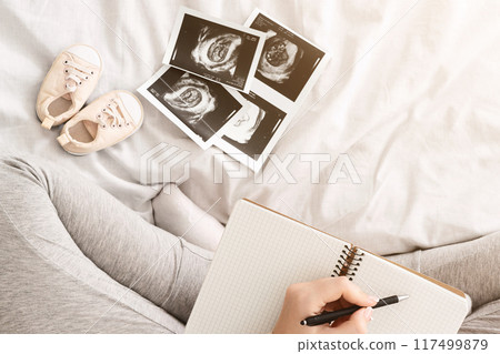 An expectant mother is sitting on a bed writing in a journal. Surrounding her are baby ultrasound images and a pair of tiny baby shoes, capturing a quiet moment of anticipation and planning 117499879