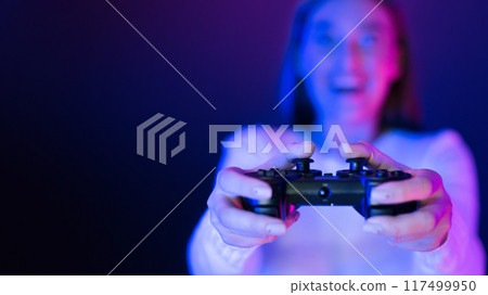 A woman is shown holding a video game controller in her hands, her fingers positioned to press the buttons. The controller is connected to a gaming console or computer, copy space A woman is shown holding a video game controller in her hands, her fingers positioned to press the buttons. The controller is connected to a gaming console or computer, copy space 117499950