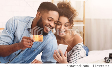 African American cheerful young couple sits closely together as they make an online purchase using a mobile phone and credit card. African American cheerful young couple sits closely together as they make an online purchase using a mobile phone and credit card. 117499953