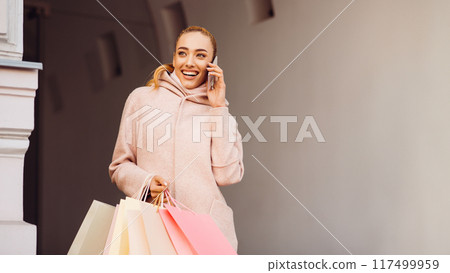 Young woman talking on phone after shopping in boutiques, walking outdoors Young woman talking on phone after shopping in boutiques, walking outdoors 117499959