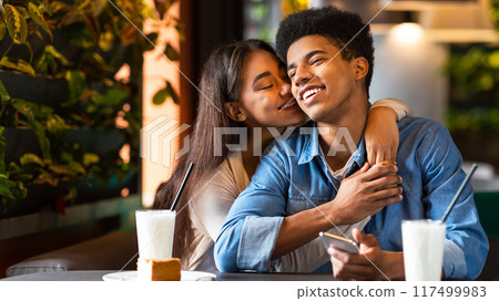African American guy and girl embrace each other warmly in a restaurant setting. They are seated at a table, surrounded by dining utensils and glasses, copy space African American guy and girl embrace each other warmly in a restaurant setting. They are seated at a table, surrounded by dining utensils and glasses, copy space 117499983