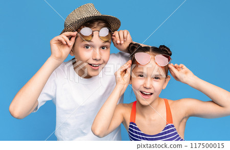 Two excited children, a boy and a girl, wearing stylish sunglasses and casual summer clothes, posing enthusiastically against a bright blue background. 117500015