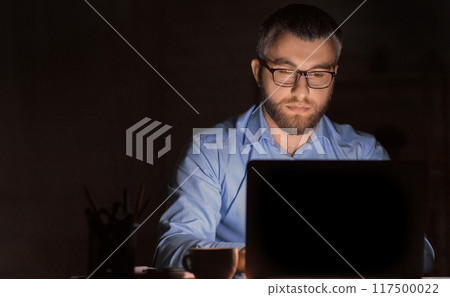 A man wearing glasses is intensely focused while working on his laptop in a dimly lit office environment during late-night hours. He appears to be deeply engaged in a task 117500022