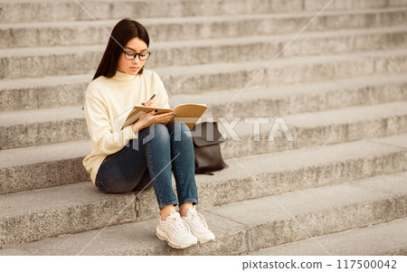 A young Asian woman with glasses, dressed in a white sweater and blue jeans, is seated on stone steps, writing in a notebook during the day. She appears focused and engaged in her task 117500042