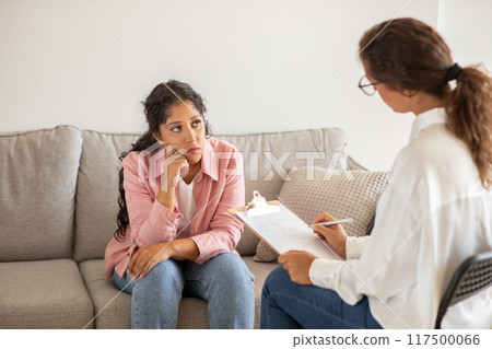 A woman sits on a gray couch in a white room while a therapist listens and takes notes. The woman looks thoughtful and concerned while the therapist appears attentive. A woman sits on a gray couch in a white room while a therapist listens and takes notes. The woman looks thoughtful and concerned while the therapist appears attentive. 117500066