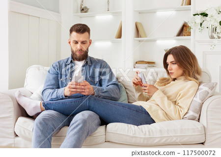 A man and woman are seated on a couch, both engrossed as they look at a cell phone screen together. A man and woman are seated on a couch, both engrossed as they look at a cell phone screen together. 117500072