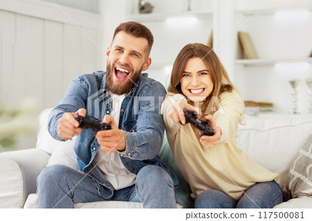 Couple sitting on a couch, engaged in a video game together. 117500081