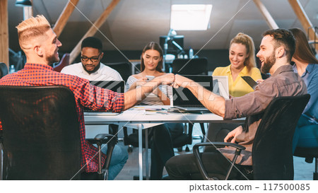 Colleagues in a loft office engage in teamwork, with two men sharing a fist bump, highlighting a positive and collaborative work environment. Colleagues in a loft office engage in teamwork, with two men sharing a fist bump, highlighting a positive and collaborative work environment. 117500085