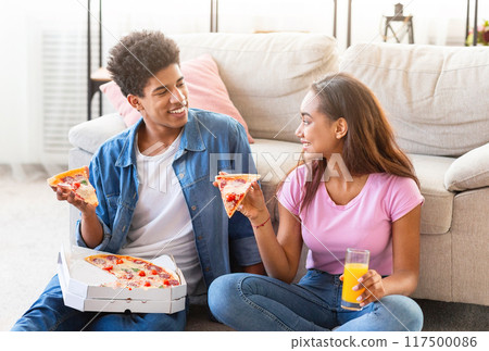 Black teen couple sits on the floor of their living room, happily eating pizza together. They are both smiling and enjoying their meal. Black teen couple sits on the floor of their living room, happily eating pizza together. They are both smiling and enjoying their meal. 117500086